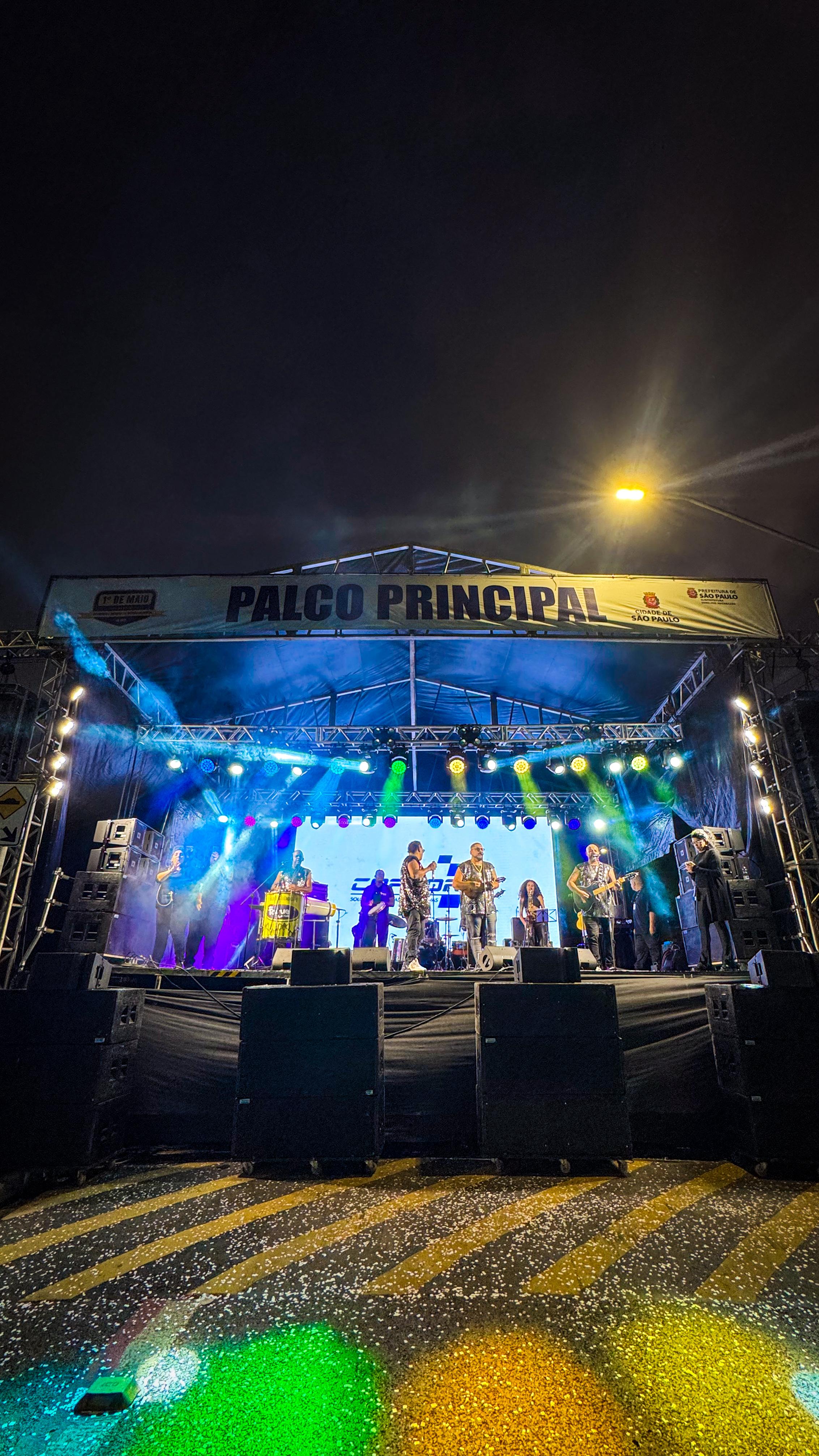 A imagem mostra um palco montado para um show noturno, iluminado por várias luzes coloridas em tons de azul, verde, roxo e amarelo que criam feixes de luz no ar. No topo do palco, uma faixa branca com a inscrição "PALCO PRINCIPAL" em letras pretas e os logotipos de "CIDADE DE SÃO PAULO" e "ERMELINO MATARAZZO E PONTE RASA" em suas extremidades. No palco, uma banda se apresenta, com vários músicos tocando instrumentos como guitarra e bateria. Na parte inferior da imagem, caixas de som pretas estão dispostas em frente ao palco. O chão, que é uma rua, reflete as luzes do palco, criando um efeito visual interessante. O céu noturno é visível acima do palco.