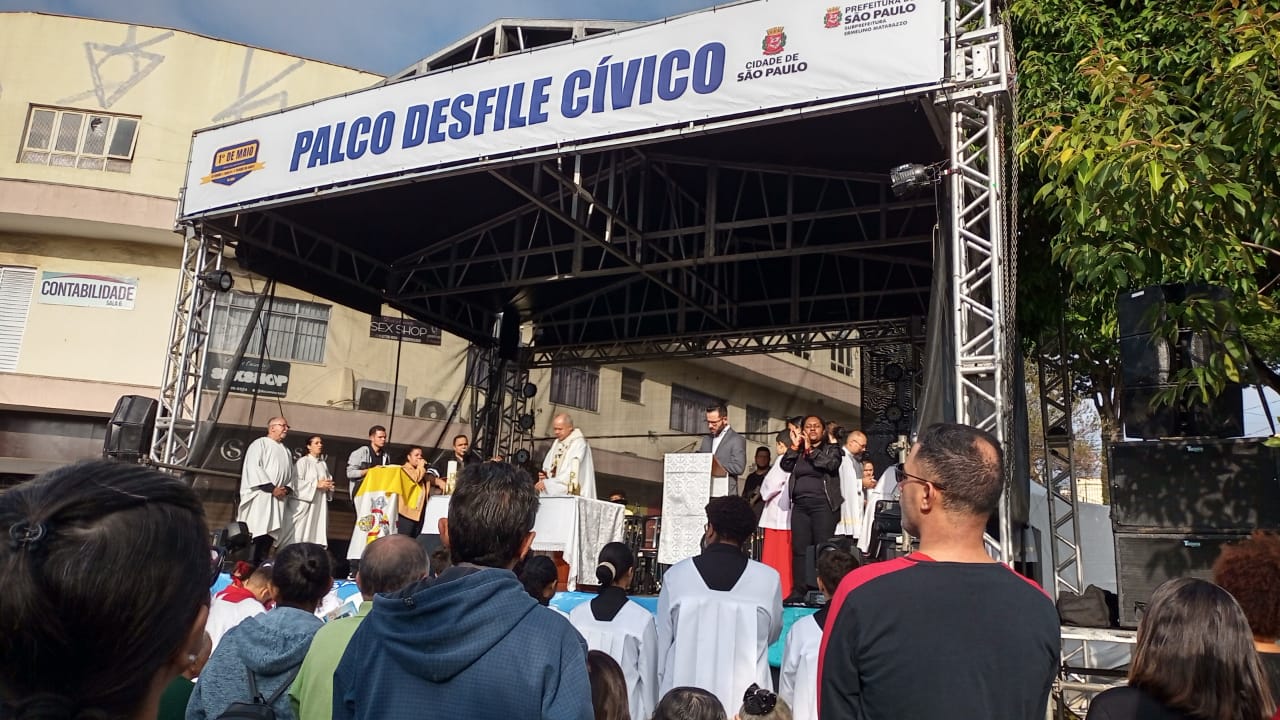 A imagem mostra um palco montado ao ar livre para um evento cívico, com uma faixa branca no topo exibindo "PALCO DESFILE CÍVICO" em letras azuis e o logotipo da "CIDADE DE SÃO PAULO". No palco, algumas pessoas vestidas com túnicas brancas e outras em roupas sociais estão reunidas, sendo que uma pessoa em destaque parece estar conduzindo uma cerimônia, possivelmente um líder religioso. Em frente ao palco, uma plateia de pessoas de costas ou de perfil assiste ao evento, algumas delas também vestindo camisetas brancas. Ao fundo, prédios urbanos e folhagens de árvores completam a cena. O céu está claro, indicando que é dia.