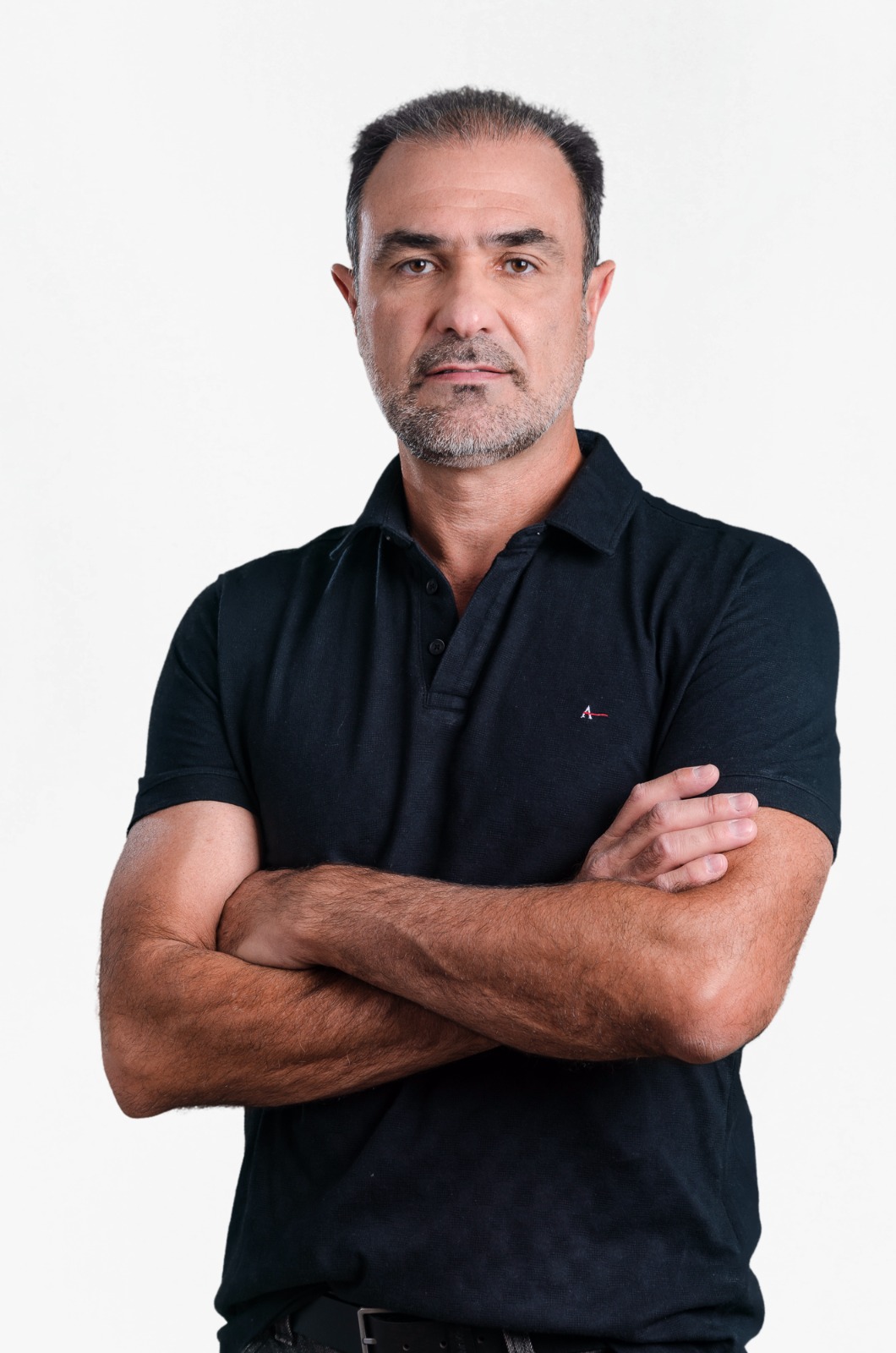 Foto em plano médio de um homem de meia-idade, com barba e cabelo escuros e curtos, vestindo uma camisa polo preta. Ele está de frente para a câmera, com uma expressão séria e os braços cruzados sobre o peito. O fundo é branco e liso. Acima da imagem, há o texto em português "Subprefeito".