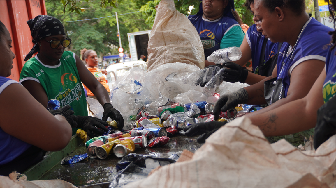 Close-up focado nas mãos de trabalhadores durante a triagem de materiais recicláveis. Diversas pessoas usando luvas pretas manipulam uma pilha de latas de alumínio amassadas e garrafas plásticas transparentes sobre uma superfície plana. Alguns vestem camisetas verdes e outros azuis, com a logomarca do "Bloco da Reciclagem". Grandes sacos de ráfia brancos são utilizados para coletar e separar os itens. A cena mostra o trabalho detalhado de separação manual em um ambiente externo arborizado.